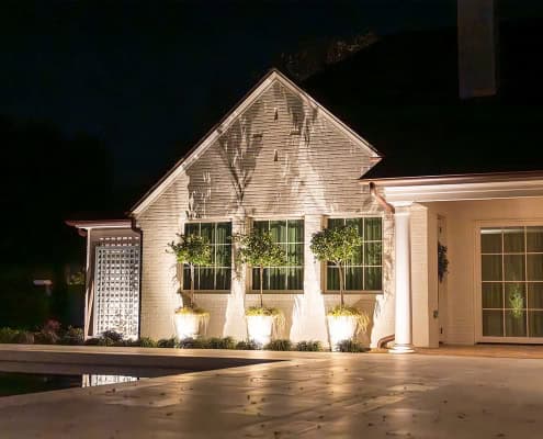 The Clover Ridge Outdoor Lighting Project Exterior night view of Clover Ridge home featuring three symmetrically arranged windows, each with a potted topiary, illuminated by landscape lighting. A pool reflects the soft glow, enhancing the elegant ambiance.