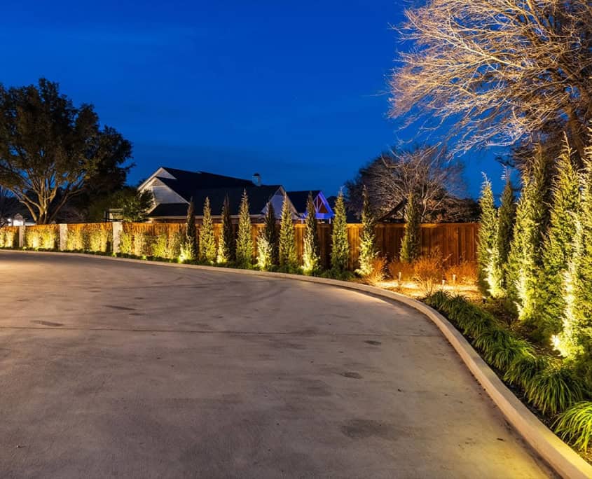 Night view of Clover Ridge landscaping, featuring illuminated trees and a curved driveway. Warm lights highlight the fence and greenery, creating an inviting, upscale ambiance.