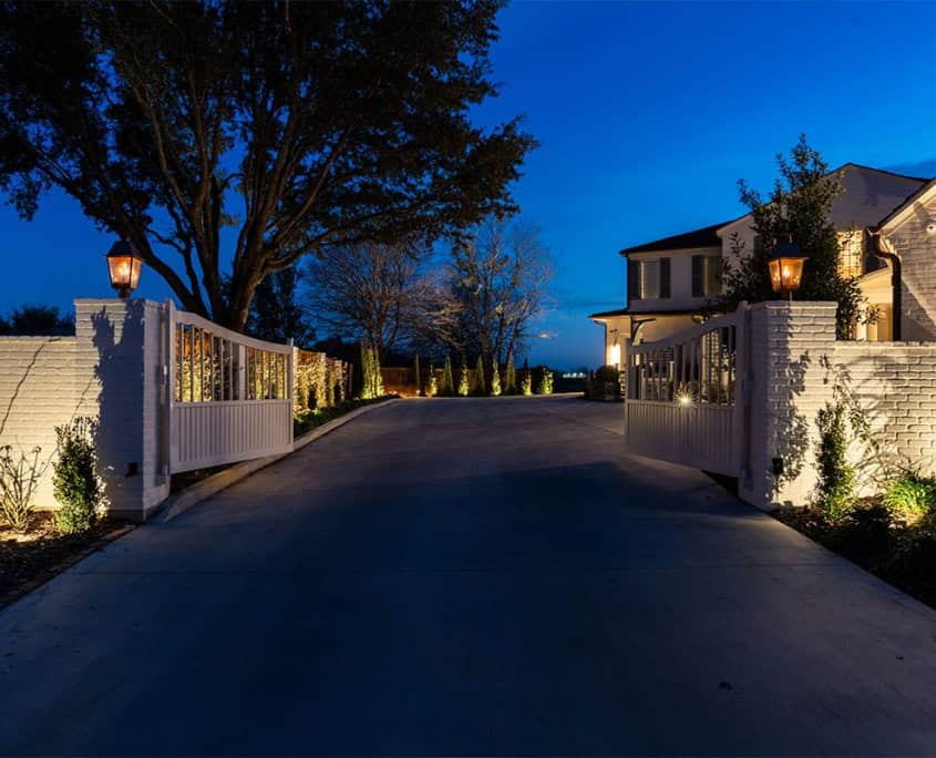 An illuminated driveway leads through white brick gates into the Clover Ridge estate at dusk. The long driveway is lined with trees and landscaping, creating an inviting entrance.