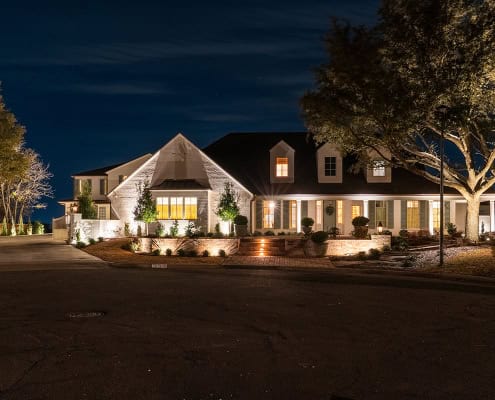 The Clover Ridge Outdoor Lighting Project A stately home in Clover Ridge glows at night, its warm interior lights spilling onto the manicured lawn and highlighting the architectural details of the facade. The dark sky provides a dramatic backdrop.