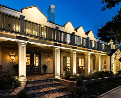 Two-story house at dusk with lit columned front porch, second-floor balcony, brick steps and landscaped flowerbeds.