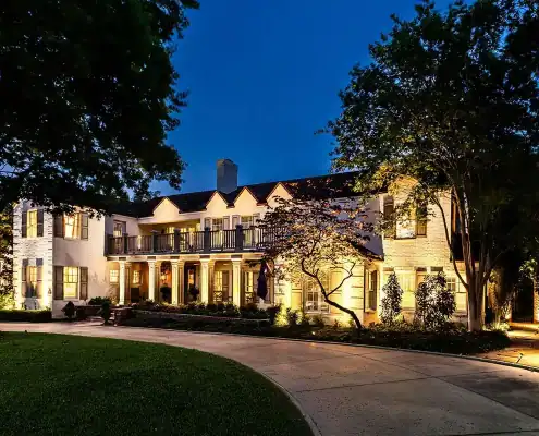 Two-story illuminated colonial-style house at dusk with front columns and balcony, curved driveway and trees in foreground.