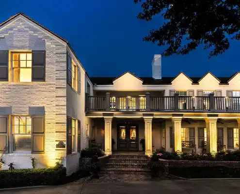 Two-story white brick house at dusk with warm lit windows, columned front porch, second-floor balcony, brick steps and low landscaping.