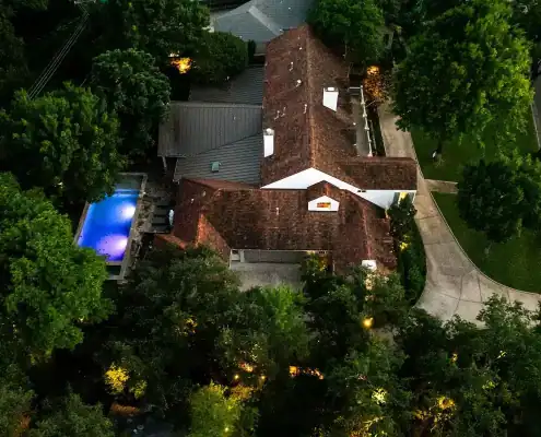 Aerial nighttime view of a large suburban house with a lit blue swimming pool, surrounding trees and a curved concrete driveway.