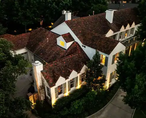 Aerial view of a large white two-story house with red tiled roof, illuminated windows and exterior lights, surrounded by trees and curved driveway.