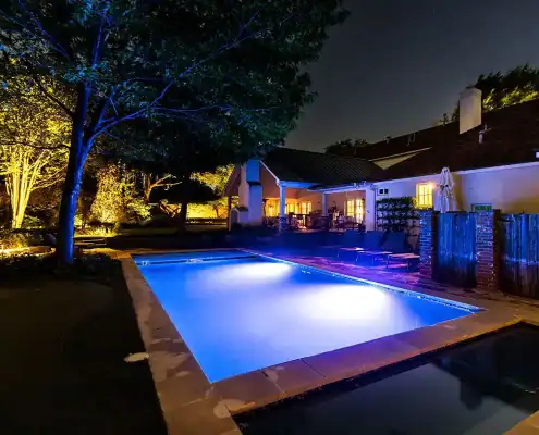 Night backyard with a glowing blue rectangular pool, patio loungers, illuminated trees and a house with warm-lit windows.