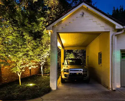 SUV parked in a covered carport beside a white brick house at night, warm lights illuminating nearby trees and a brick garden wall.