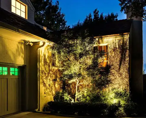 Night view of a house exterior with a lit tree and shrubs casting shadows on a brick wall, illuminated windows and garage door with green panes.