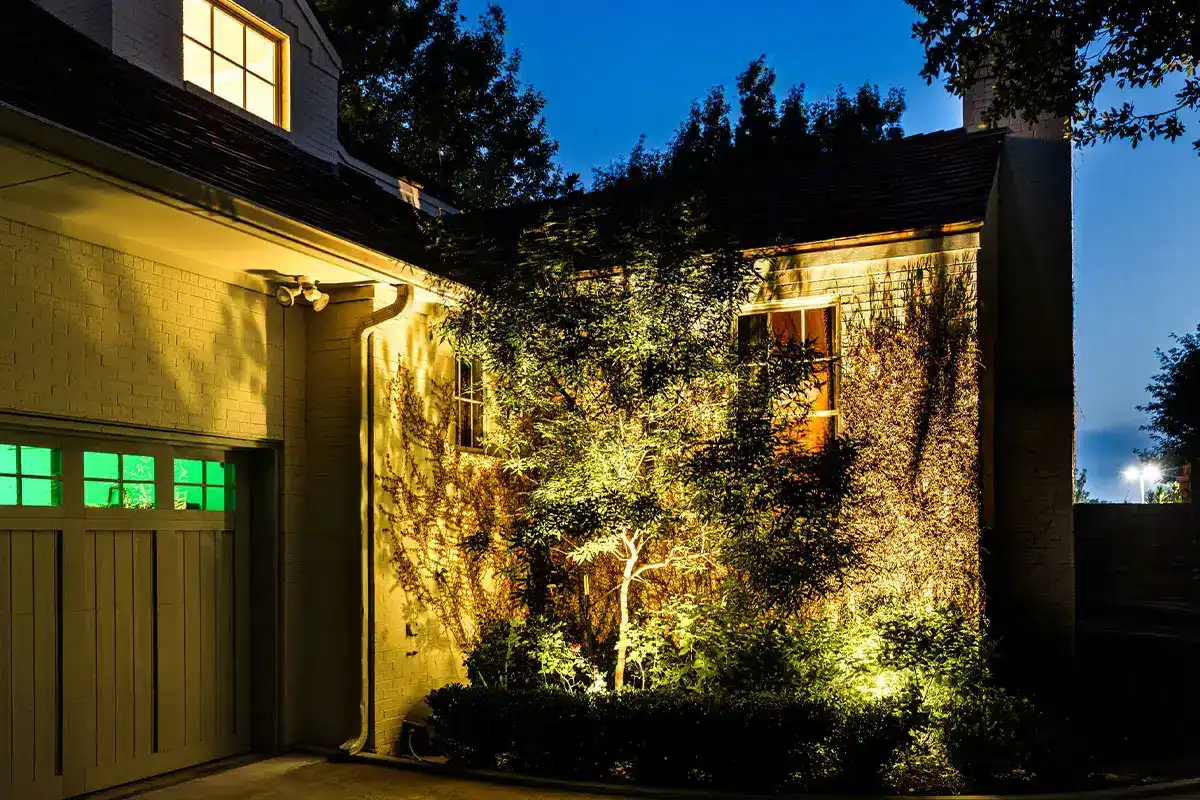 Night view of a house exterior with a lit tree and shrubs casting shadows on a brick wall, illuminated windows and garage door with green panes.