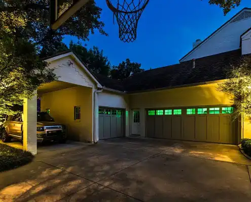 Dusk driveway with SUV in a covered carport, three-car garage with green-lit windows, illuminated trees and a basketball hoop overhead.