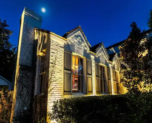 Night view of a white brick house with gabled windows glowing from interior light, a tall chimney, moonlit deep-blue sky, and surrounding trees and shrubs.