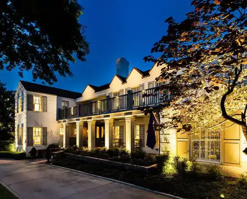 Two-story white house at dusk with illuminated porch columns, upper balcony, lit windows, driveway and a tree in foreground.