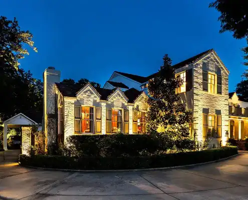 Large two-story illuminated brick house at dusk with lit windows, manicured shrubs, curved driveway and a covered carport to the left.