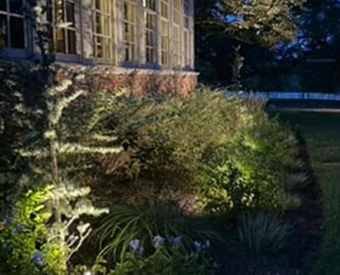 Outdoor lighting illuminates a stone house and lush garden at night. The soft glow highlights the textures of the stonework and the greenery, creating a warm, inviting ambiance.