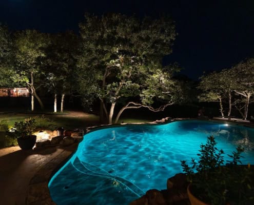 Nighttime view of a turquoise-lit pool surrounded by lush trees illuminated by landscape lighting. The scene evokes a sense of poolside serenity under a moonlit canopy design.