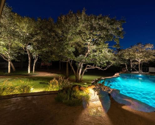 Night view of a backyard pool illuminated with turquoise lighting, reflecting the moonlit canopy design of trees spotlighted around the yard. Patio and lush landscaping surround the pool.