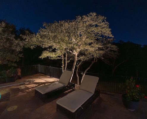 Two lounge chairs sit poolside under a tree illuminated by outdoor lighting at night, creating a serene moonlit canopy design. The pool glows blue, and stars are visible in the dark sky.