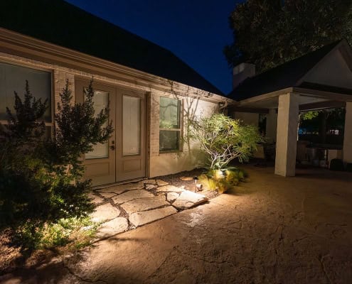 Night view of poolside canopy design, illuminated by landscape lighting. Stone patio leads to French doors, with lush greenery adding to the serene ambiance.
