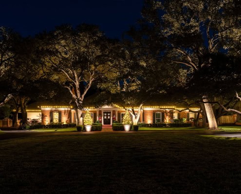 A stately home in Ridgeview at night, illuminated by warm outdoor lighting. Trees frame the house, their branches also lit, creating a welcoming and festive ambiance.