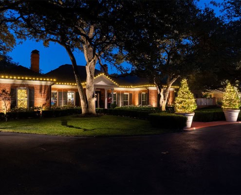 Brick Ridgeview home at night, illuminated with warm Christmas lights along the roofline. Two potted Christmas trees flank the driveway, adding festive cheer.