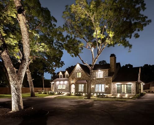 Stone mansion at night, exterior lights and lit windows, tall trees in front and curved driveway.