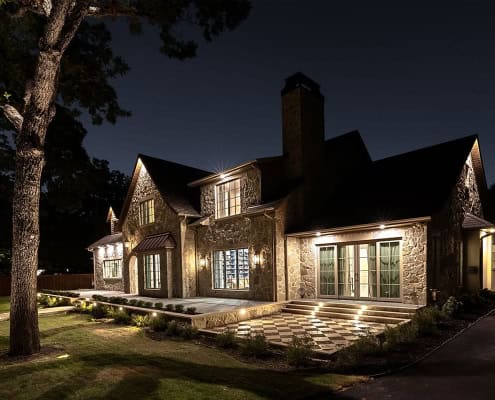 Stone two-story house at night with warm exterior lighting, illuminated patio and steps, large tree and manicured lawn in foreground.