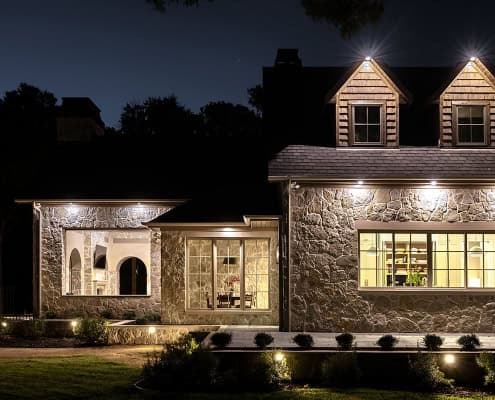Night view of a stone house exterior with illuminated windows, dormers and porch lights, interior dining and kitchen visible through large windows.