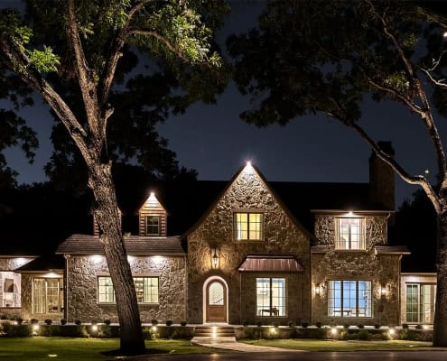 Stone two-story house at night with warm interior and exterior lights, framed by two large trees and a lit front walkway.