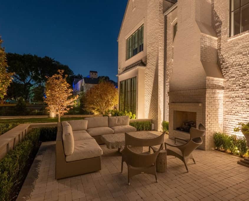 Outdoor patio at dusk featuring a cream-colored sectional sofa, wicker chairs, and a stone-topped table, illuminated by landscape lighting. A brick fireplace and manicured hedges add to the elegant ambiance of The Desco Project design.