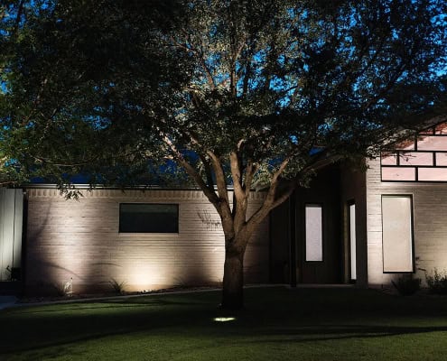 Night view of a modern home, part of The Falcon Project, with landscape lighting illuminating the brick facade and mature trees, creating a warm and inviting ambiance.