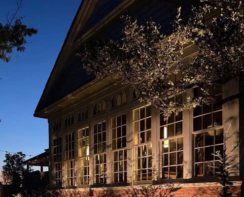 The Farm Project: A building with large windows illuminated at dusk. Warm light spills from the windows, contrasting with the deep blue sky.
