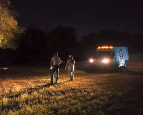 Two people stand in a field at night, illuminated by a van with flashing lights. The Goldfawn Ranch Project may involve emergency response or late-night work.