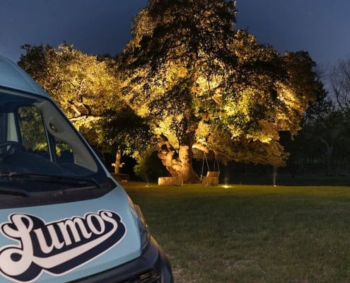 Lumos lighting van parked near a grand, illuminated oak tree at Goldfawn Ranch. The tree is bathed in golden light, creating a magical nighttime scene. "Lumos" logo visible on the van.