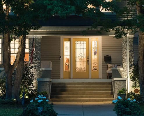 Inviting home exterior at dusk. Warm light spills from the yellow front door and windows of this charming house, nestled amidst lush trees and landscaping. Steps lead to the porch, creating a welcoming scene.