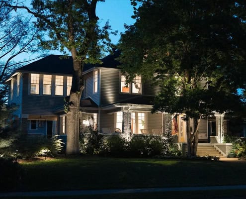 Exterior of a beautifully lit two-story home at dusk, showcasing the finished "Maple Project" with warm light spilling from windows and highlighting the manicured lawn and mature trees.