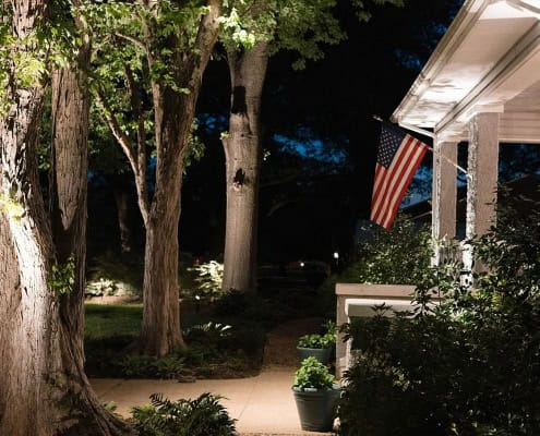 Night view of a home illuminated by landscape lighting, showcasing the "Maple Project". An American flag hangs from the porch, with trees and foliage bathed in warm light along the walkway.
