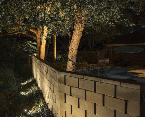 Nighttime view of the Woodbriar Project's landscape lighting, showcasing a retaining wall softly lit by in-ground lights, with trees illuminated from below, and a pool area in the background.