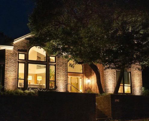 Elegant brick home at night, part of The Woodbriar Project, with illuminated arched windows and lush tree in the front yard. Warm light spills from the interior, creating a welcoming ambiance.