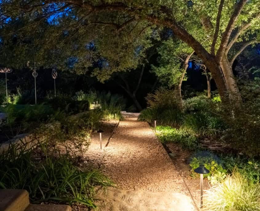 Softly illuminated gravel pathway with trees on the right and foilage on both sides of the walkpath.