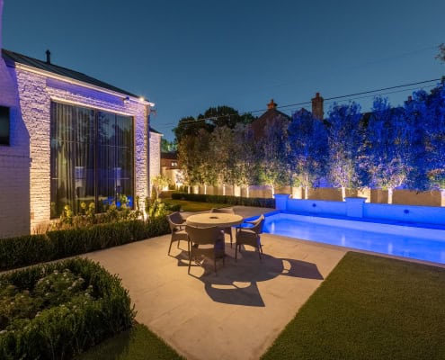 The Desco Outdoor Lighting Project Twilight view of The Desco Project's backyard featuring a rectangular pool illuminated with blue lights, manicured lawn, and modern white brick home with a tall chimney.