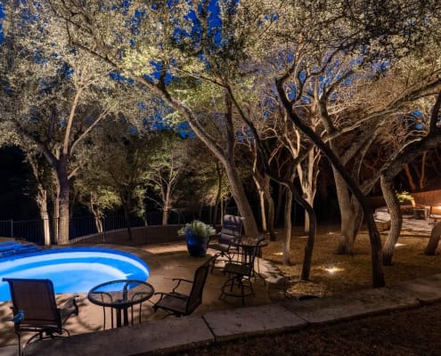 Outdoor lighting softly illuminating trees around a patio with a fire pit on the right side and an illuminated pool with patio chairs surrounding it on the left.